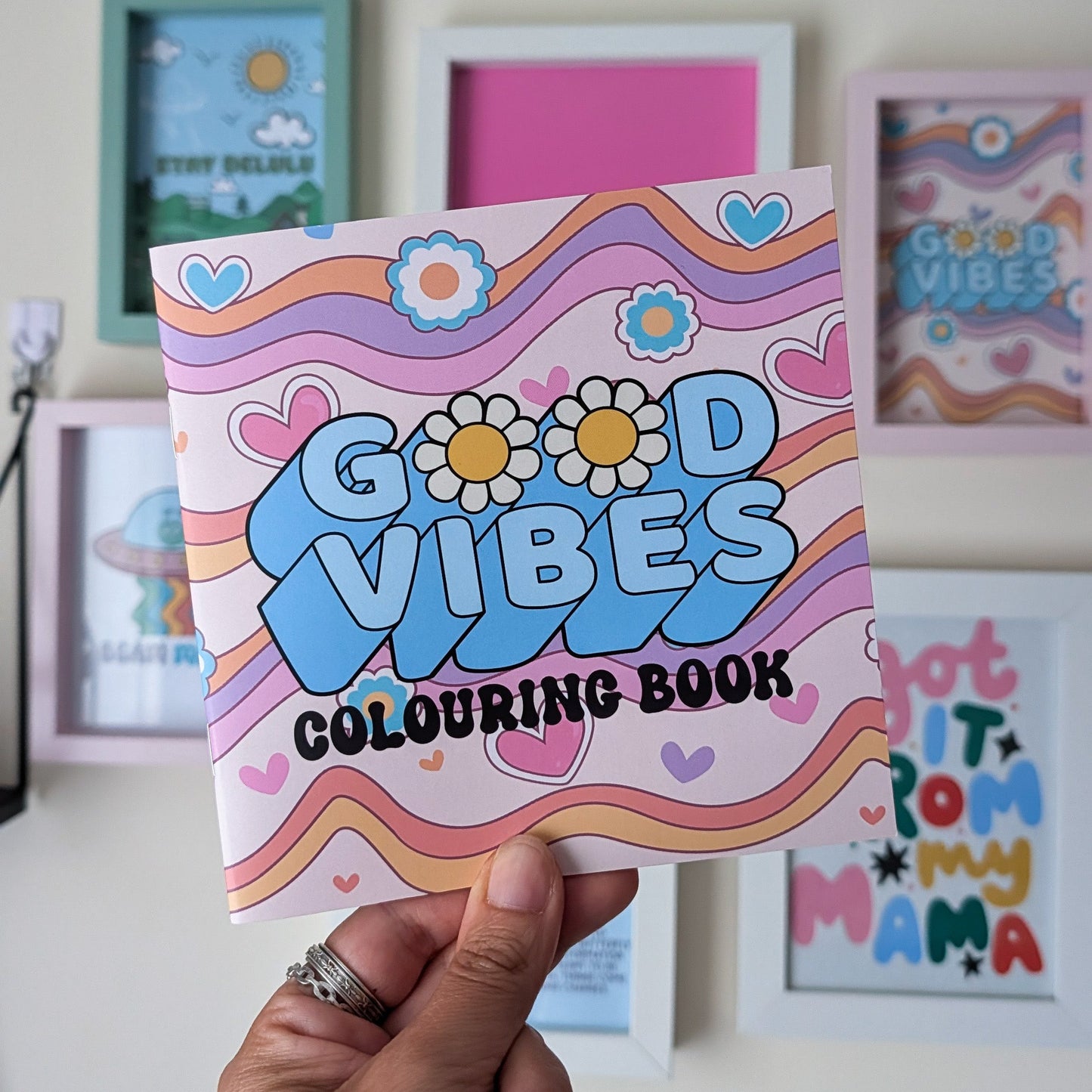 A hand holds a colorful 'Good Vibes Colouring Book' in front of a wall with framed art. The book cover features retro wavy stripes in pink, orange, yellow, and blue, with the words 'GOOD VIBES COLOURING BOOK' in playful lettering.