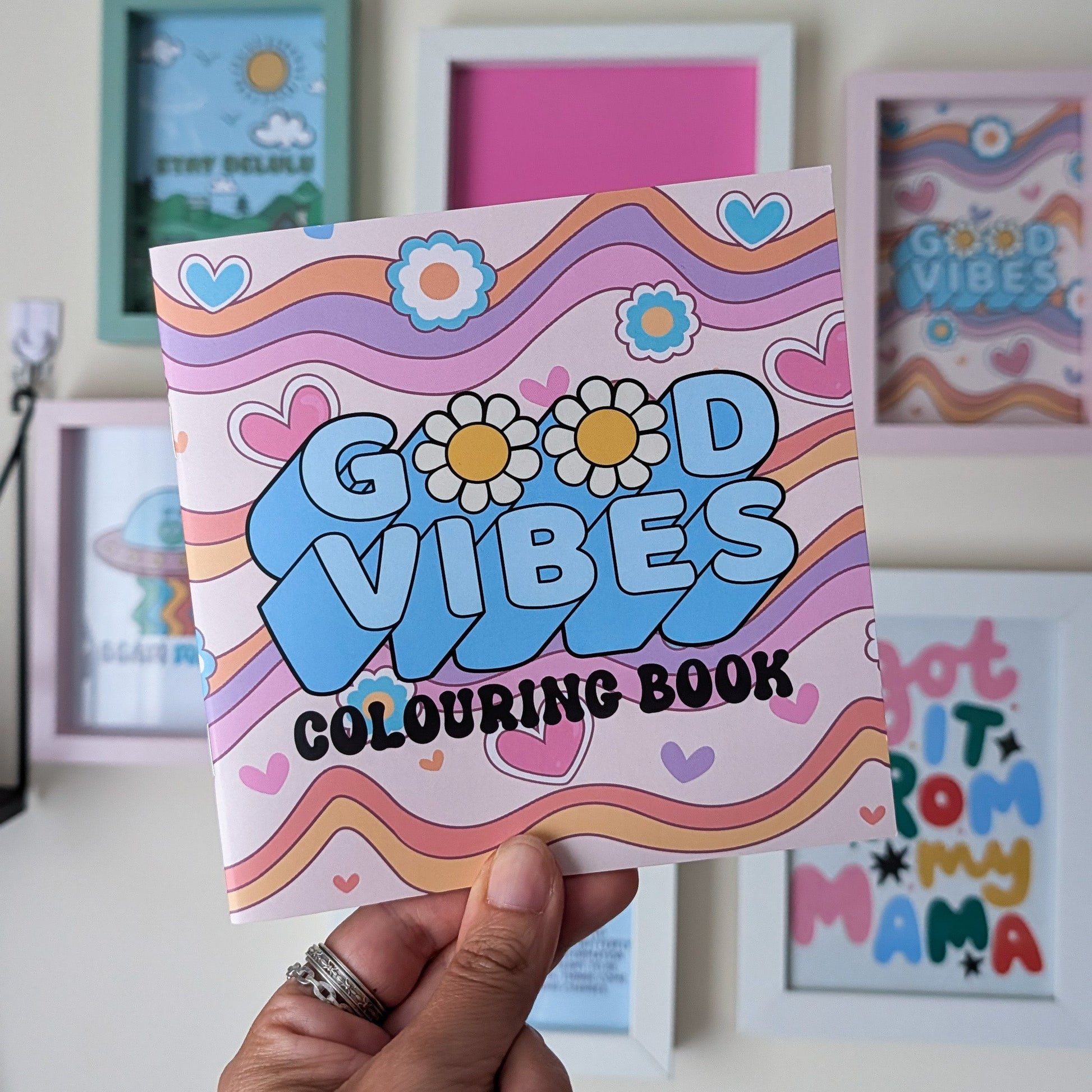 A hand holds a colorful 'Good Vibes Colouring Book' in front of a wall with framed art. The book cover features retro wavy stripes in pink, orange, yellow, and blue, with the words 'GOOD VIBES COLOURING BOOK' in playful lettering.